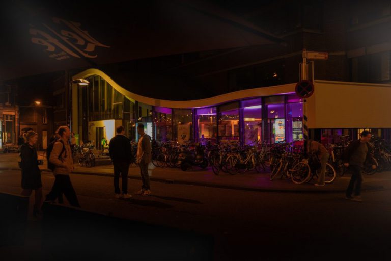 Exterieur van het Festivalhuis tijdens de Kunstnacht. Buiten is het donker. Er komen paarse lichten door de ramen van het gebouw. Verschillende groepjes mensen lopen over de straat voor het pand en zijn met elkaar in gesprek.